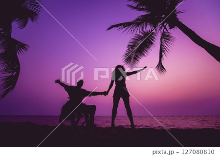 Disabled man in a wheelchair with his wife on the beach. Silhouettes at sunset 127080810