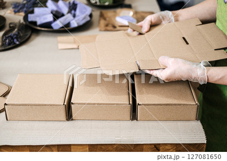 Person folds kraft boxes at a table near handmade soaps and eco filler. Scalable packaging, modular box system, ecommerce adaptability, minimal waste design 127081650