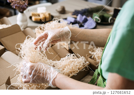 Gloved hands arrange green and lavender soaps with dried flowers in kraft packaging. Handmade business, eco-friendly packaging, natural skincare, artisan presentation 127081652