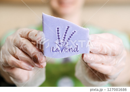 Smiling person in gloves holds up a stamped lavender soap bar for the camera. Lavender soap, handmade branding, aromatherapeutic skincare, plant-based wellness Smiling person in gloves holds up a stamped lavender soap bar for the camera. Lavender soap, handmade branding, aromatherapeutic skincare, plant-based wellness 127081686
