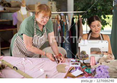 Mature woman cutting fabric next to female seamstress 127086240