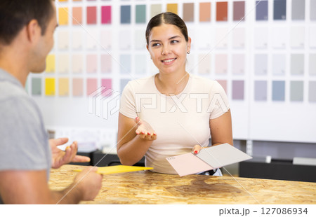 Female saleswoman of paintwork material shop advises man buyer with samples and product catalog 127086934