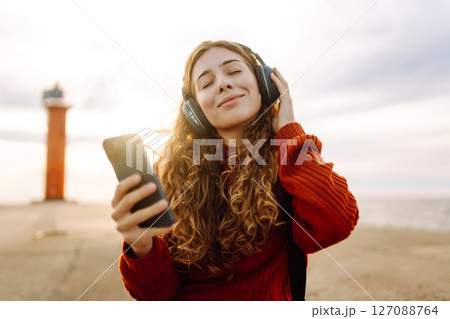 Happy woman listening nice music by the waterfront while wearing headphones by a lighthouse. Happy woman listening nice music by the waterfront while wearing headphones by a lighthouse. 127088764