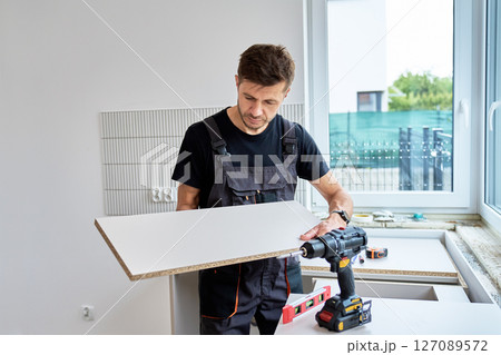Man in uniform measures cabinet panel during kitchen furniture installation. Renovation process in new apartment Man in uniform measures cabinet panel during kitchen furniture installation. Renovation process in new apartment 127089572