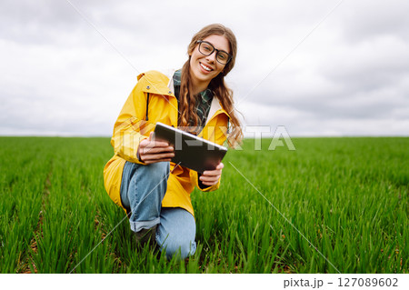 Young farmer woman in a green field, intently using a tablet. She wears a yellow jacket and glasses Young farmer woman in a green field, intently using a tablet. She wears a yellow jacket and glasses 127089602