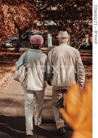 Elderly couple walking in autumn park. Elderly couple in light jackets walks together through peaceful autumn park with golden foliage. Concept of companionship, aging with love, autumn stroll Elderly couple walking in autumn park. Elderly couple in light jackets walks together through peaceful autumn park with golden foliage. Concept of companionship, aging with love, autumn stroll 127089622