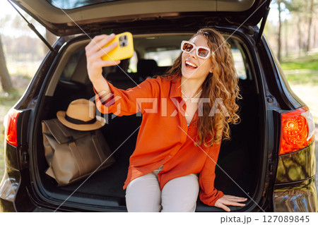 Happy woman sits on the trunk of her car and taking a selfie. Selfie while traveling by car. 127089845