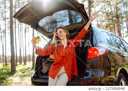Happy woman sits on the trunk of her car and taking a selfie. Selfie while traveling by car. 127089846