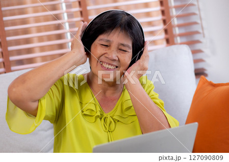 Happy senior asian woman wearing headphones listening music while sitting on sofa with fun. Happy senior asian woman wearing headphones listening music while sitting on sofa with fun. 127090809