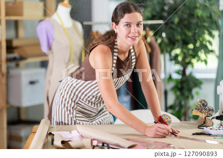 Female dressmaker draws sketch of future clothing on paper using patterns in sewing workshop Female dressmaker draws sketch of future clothing on paper using patterns in sewing workshop 127090893