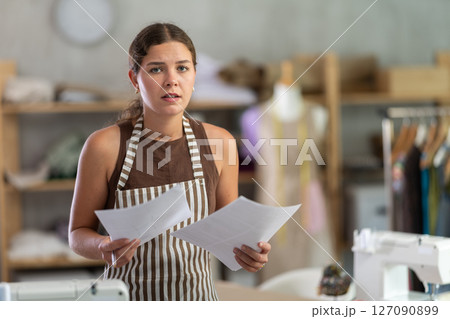 Sad seamstress girl reading financial document in sewing studio interior 127090899