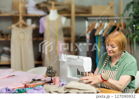 Mature female seamstress is sitting near sewing machine and create new dress in workshop Mature female seamstress is sitting near sewing machine and create new dress in workshop 127090932