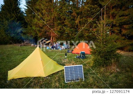 Solar panel in grassy field with tents in background highlights eco-friendly camping setup. Scene includes campers, emphasizing integration of renewable energy in outdoor activities. Solar panel in grassy field with tents in background highlights eco-friendly camping setup. Scene includes campers, emphasizing integration of renewable energy in outdoor activities. 127091174
