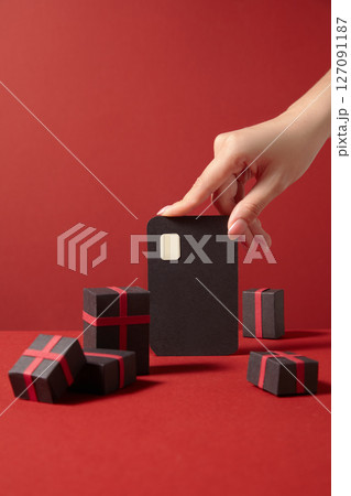 Hand holding a paper cutout ATM card among gift boxes on a red background, representing mobile apps for flash sales and shopping events 127091187