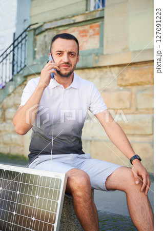Handsome man using smartphone connected to photovoltaic solar panel. Integration of sustainable renewable energy into everyday life, demonstrating practical use of solar power for charging devices. Handsome man using smartphone connected to photovoltaic solar panel. Integration of sustainable renewable energy into everyday life, demonstrating practical use of solar power for charging devices. 127091223
