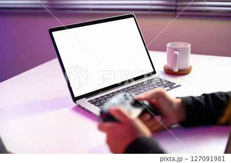 Hands holding game controller in front of a blank laptop screen with purple ambient light 127091981