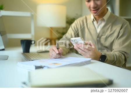 Smiling man checking his smartphone while writing notes on financial documents 127092007