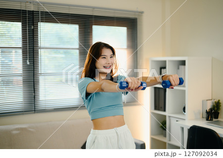 Cheerful young woman exercises with small dumbbells in her living room 127092034