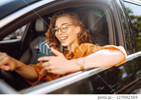 Happy woman in the car in the driver's seat looks at the smartphone. Business, technology concept. 127092369