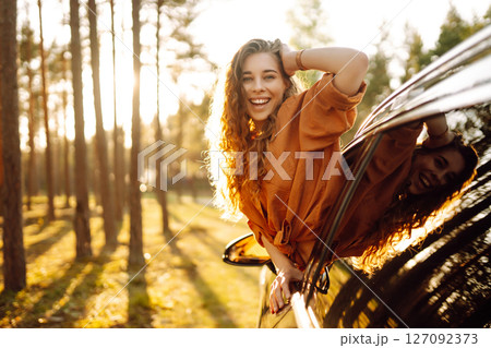 A young woman feels freedom leaning out of a car window in a sunny forest. Travel concept. A young woman feels freedom leaning out of a car window in a sunny forest. Travel concept. 127092373