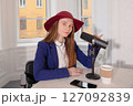 A young woman in a hat is recording a podcast. The woman is speaking into a table microphone. 127092839