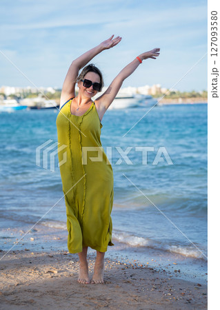 Woman in yellow dress stands against blue sea and beach waving her hands Woman in yellow dress stands against blue sea and beach waving her hands 127093580