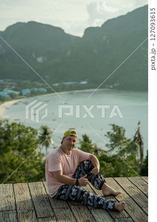 A guy in a pink wet T-shirt and Thai pants sits on a terrace overlooking an island at sunset A guy in a pink wet T-shirt and Thai pants sits on a terrace overlooking an island at sunset 127093615