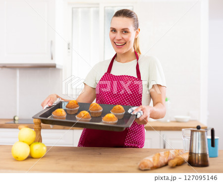 Smiling woman in apron standing in home kitchen with tray of cupcakes Smiling woman in apron standing in home kitchen with tray of cupcakes 127095146