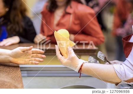Ice cream seller giving ice cream cone to customer. Vendor in glove handing yellow ice cream cone to customer across counter. Concept of ice cream, food service, organic summer refreshment Ice cream seller giving ice cream cone to customer. Vendor in glove handing yellow ice cream cone to customer across counter. Concept of ice cream, food service, organic summer refreshment 127096986