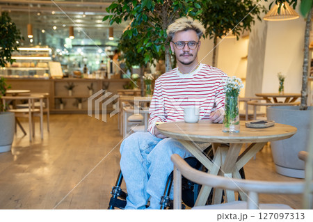 Portrait of thoughtful, calm, serene man in wheelchair enjoys coffee in cafe, sits alone at table 127097313
