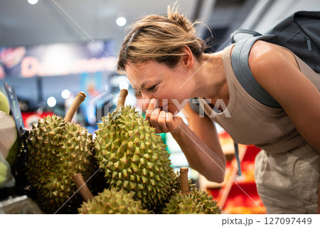 Confused woman sniffs durian with disgust on stall, facial expression with rejection of strong scent Confused woman sniffs durian with disgust on stall, facial expression with rejection of strong scent 127097449