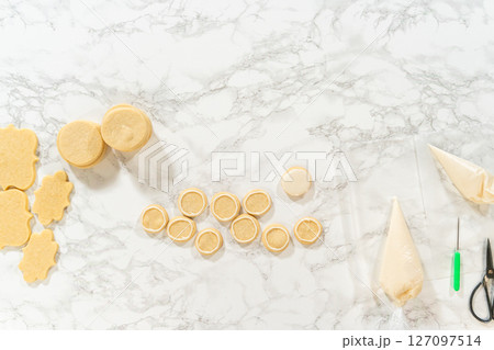 Freshly baked round sugar cookies arranged on a marble surface, prepped for royal icing decoration. A piping bag, scribe tool, and cookie scraps are visible, showing the decorating process. Freshly baked round sugar cookies arranged on a marble surface, prepped for royal icing decoration. A piping bag, scribe tool, and cookie scraps are visible, showing the decorating process. 127097514