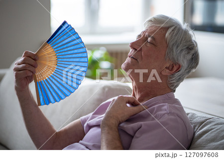 Heat can be deadly for seniors. Sweltering pensioner with fan struggling with summer heat indoors 127097608