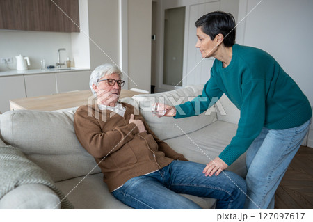 Caring wife helps elderly suffering husband clutching chest sitting on sofa, offering glass of water 127097617