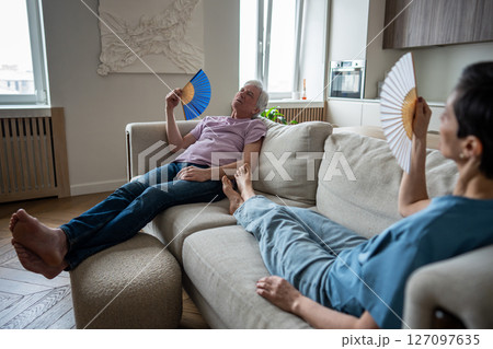 Stressed elderly couple suffering from heat, fanning on sofa unable to function. Seniors struggle 127097635