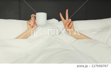 Cheerful lazy woman waking up after sleeping lying in bed holding cup in her hands asks for coffee Cheerful lazy woman waking up after sleeping lying in bed holding cup in her hands asks for coffee 127098767