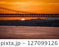 Beautiful sunset over 25 de Abril bridge in silhouette and Tagus river with sailboats drifting below and Lisbon skyline in summer haze in the background. Orange sky, clouds and sun, tranquil waters 127099126