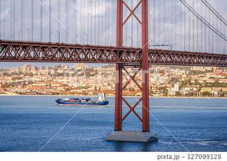 Colorful landscape with blue container ship on Tagus river under iconic 25 de Abril or 25th April bridge, Lisbon, Portugal. Famous ironwork of red suspension bridge, vibrant cityscape, urban 127099128