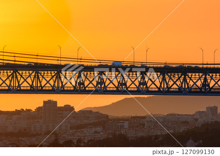 Beautiful view of golden sunset and suspension city bridge with its steel structure and cables, dramatic silhouettes of vehicles in motion blur against bright orange sky. Distant hills and urban 127099130