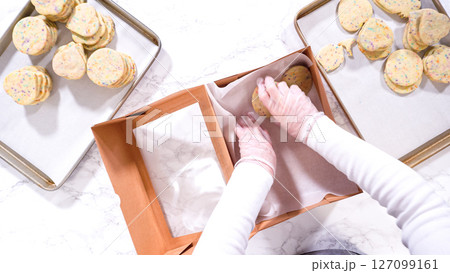 Flat lay. With precision, the woman is carefully arranging the sugar cookies, filled with dough-mixed sprinkles, into a rustic brown paper box. Flat lay. With precision, the woman is carefully arranging the sugar cookies, filled with dough-mixed sprinkles, into a rustic brown paper box. 127099161