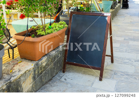 Blank wooden chalkboard stands beside flower planter on stone walkway. Outdoor signage, seasonal menu display, and cozy cafe atmosphere. Copy space Blank wooden chalkboard stands beside flower planter on stone walkway. Outdoor signage, seasonal menu display, and cozy cafe atmosphere. Copy space 127099248