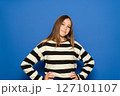 Pretty Hispanic teenage girl wearing a black and white striped sweater with her arms akimbo, waiting for something, isolated on a plain blue studio background. Lifestyle concept. 127101107