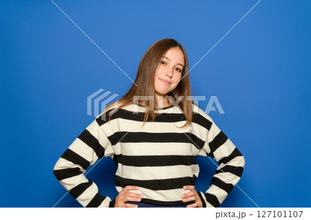 Pretty Hispanic teenage girl wearing a black and white striped sweater with her arms akimbo, waiting for something, isolated on a plain blue studio background. Lifestyle concept. Pretty Hispanic teenage girl wearing a black and white striped sweater with her arms akimbo, waiting for something, isolated on a plain blue studio background. Lifestyle concept. 127101107