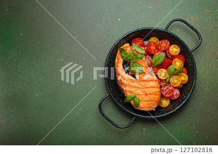Grilled salmon steak with cherry tomatoes and basil leaves in black pan with cutlery top view on dark green background, space for text Grilled salmon steak with cherry tomatoes and basil leaves in black pan with cutlery top view on dark green background, space for text 127102816