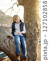 A cute preteen boy is sitting on a branch of a large sycamore tree 127102858