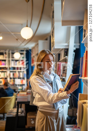 Focused middle aged stylish woman choosing book in bookstore, standing near window reads content. 127104258