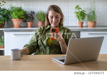 Joyful woman taking break in kitchen, looking on smartphone chatting, scrolling web holding cup tea Joyful woman taking break in kitchen, looking on smartphone chatting, scrolling web holding cup tea 127104271