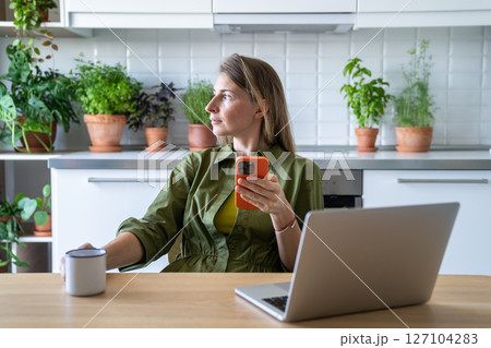 Thoughtful woman sits in kitchen with cup holding smartphone immersed daydreaming looking to window. Thoughtful woman sits in kitchen with cup holding smartphone immersed daydreaming looking to window. 127104283