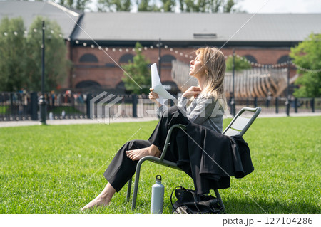 Uncomfortable woman fanning with sheets of paper like fan while sitting on chair outside on lawn. 127104286