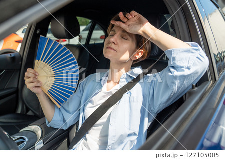 Exhausted middle aged woman drives car waves blue fan suffers from stuffiness in traffic jam. Exhausted middle aged woman drives car waves blue fan suffers from stuffiness in traffic jam. 127105005